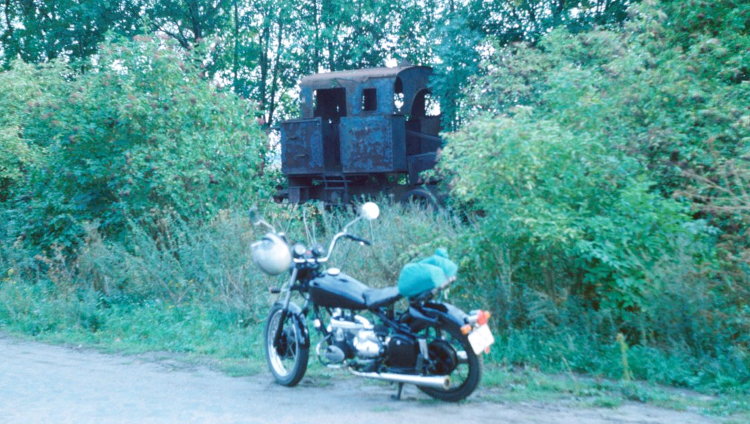 das Motorrad von links vor einem im Gebüsch halb versteckten Dampfloktorso